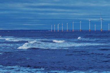 Açıklardaki rüzgar türbinleri mavi bir gökyüzünün altında okyanusta bir çizgide dururlar. Dalgalar ön planda görünür. Uzak kıyı şeridi İngiltere, Redcar 'da belli belirsiz görünür..