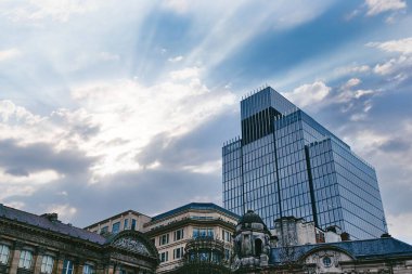 Modern bir cam gökdelen, parçalı bulutlu bir gökyüzünün altındaki eski ve süslü taş binalara karşı birleşerek gökyüzüne hükmediyor. Birmingham, İngiltere 'de güneş ışınları bulutları delip geçer..