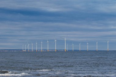 Açıklardaki rüzgar türbinleri bulutlu bir gökyüzünün altında bir su kütlesi boyunca bir hat halinde ilerler. Sahne sakin ve uzak bir kıyı şeridi Redcar, İngiltere 'de görülebiliyor..