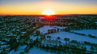 Gün batımında karla kaplı bir kasabanın havadan görünüşü. Evler, ağaçlar ve bir park parlak turuncu ve mavi gökyüzünün altında görülebilir. Güneş Harrogate, İngiltere 'de kasabanın tam arkasında batıyor..