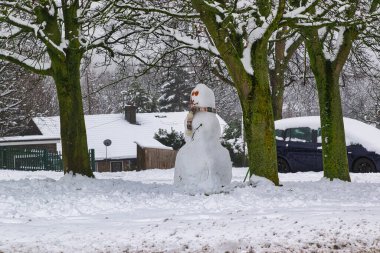 Eşarplı bir kardan adam karla kaplı iki ağacın arasında, karlı bir arazide duruyor. Harrogate, İngiltere 'de kar kaplı bir ev ve araba arka planda görünür..