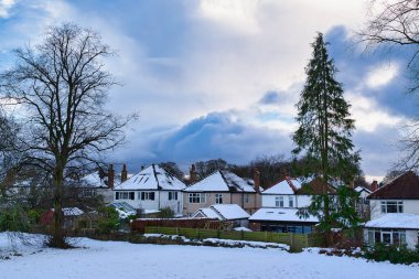 Parçalı bulutlu bir gökyüzünün altında karla kaplı banliyö evleri. Ön planda ve arka planda çıplak ağaçlar ve Evergreen 'ler görülebilir. İngiltere 'nin Harrogate kentindeki karla kaplı bir tarladan evleri ayıran tahta bir çit var..