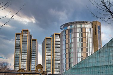 Bulutlu bir gökyüzüne karşı, kıvrımlı bir Indigo Oteli de dahil olmak üzere üç modern gökdelen sergilenmektedir. Otelin etrafını saran iki dikdörtgen bina, bir altın ve bir bronzlaşma. Manchester, U 'da önplanda cam önlü bir yapının bir kısmı görülebilir.