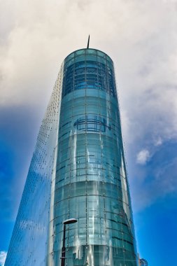 Ağırlıklı olarak deniz mavisi ve şeffaf modern cam bir binanın alçak açılı görüntüsü silindirik merkezi bir kule bölümü ve düz, panelli bir dış yüzeyi gösteriyor. Manchester, İngiltere 'de gökyüzü kısmen bulutlu..