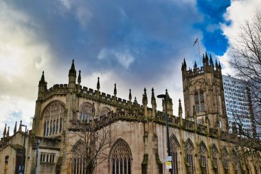 Gotik mimari ile taştan kilise cephesi, uzun merkezi bir kule ve sayısız kemerli pencereler içeriyor. Modern bir bina kısmen arka planda, Manchester, İngiltere 'de bulutlu bir gökyüzünün altında görülebilir..