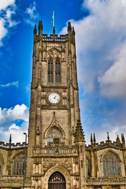 Bir saati, karmaşık oymaları olan uzun taş bir kilise kulesi ve Gotik mimari parçalı bulutlu mavi gökyüzüne karşı imaja hükmediyor. Binanın ön cephesinde Manchester, İngiltere 'de ayrıntılı taş işçiliği ve kemerli pencereler sergileniyor..
