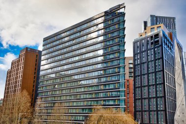 Ağırlıklı olarak koyu cam ve açılı olan bir CitySuites binası, Manchester, İngiltere 'de parçalı bulutlu bir gökyüzünün altında bronz renkli bir bina da dahil olmak üzere diğer modern gökdelenlerin arasında belirgin bir şekilde duruyor..