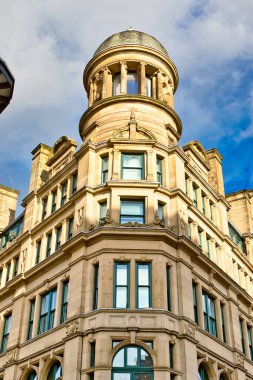 Çok sayıda çok camlı penceresi ve karmaşık mimari ayrıntıları olan, bir kubbe ile kaplanmış, bronz renkli bir taş bina. Bina Manchester, İngiltere 'de parçalı bulutlu bir gökyüzüne karşı güneş ışığıyla aydınlanıyor..