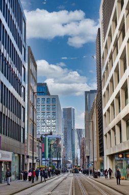 Şehir caddesinde modern binalar, yayalar ve tramvay raylarının yer aldığı bir sahne. Mimari esasen cam ve betondan oluşmaktadır ve çeşitli stiller içerir. Gökyüzü parçalı bulutlu ve Manchester, İngiltere 'deki görüntüye derinlik katıyor..