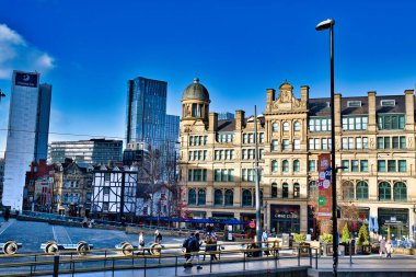 Manchester, İngiltere 'deki modern ve tarihi binalar, açık mavi gökyüzünün altında. Dekoratif detaylara sahip büyük, bronz renkli bir bina, arka planda modern gökdelenler ve Manchester, İngiltere 'de yayalar ile orta sahada egemendir.