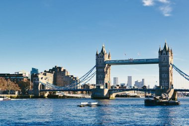 İngiltere, Londra 'daki Tower Bridge, açık mavi gökyüzünün altında Thames Nehri' ni kaplar. Binalar nehir kenarına dizilir ve bir römorkör suda görülebilir..