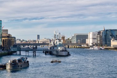 Londra 'daki Thames Nehri manzarası, rıhtımda bir savaş gemisi, bir mavna, nehir kıyısı boyunca birkaç bina ve arka planda bir köprü. Gökyüzü parçalı bulutlu.