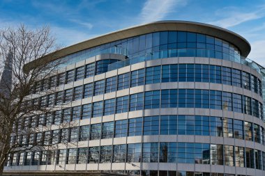 Kıvrımlı cepheli modern çok katlı bina, mavi gökyüzü ve ağaçları yansıtan sayısız pencere, kısmen görünür bir gökdelenin ve Londra, İngiltere 'deki mavi gökyüzünün zeminine karşı..