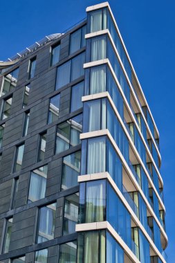 Modern building with dark gray facade and large, blue-tinted windows.  Corner view showcases multiple floors and balconies.  Bright blue sky provides contrast  in London, UK. clipart