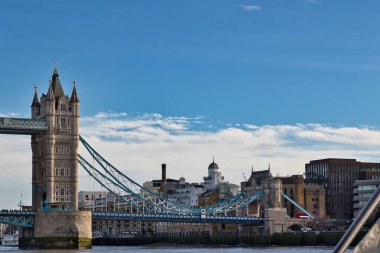 Londra 'daki Tower Bridge, İngiltere, nehirden izleniyor. Köprü ve bitişik binaların üzerinde parçalı bulutlu mavi bir gökyüzü görülebilir. Köprünün deniz mavisi süspansiyon kabloları ve taş kuleleri ön planda..