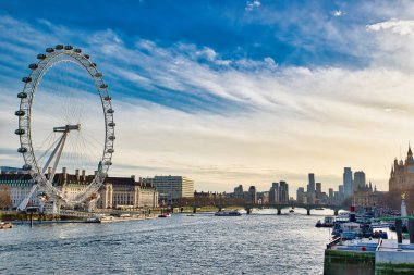 London Skyline 'da London Eye, Thames Nehri boyunca binalar, birkaç köprü ve tekneler var. Parçalı bulutlu bir gökyüzü mavi ve beyaz karışımını gösterir.