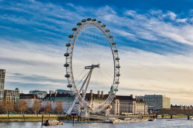 Londra Göz Dönme dolabı parçalı bulutlu mavi gökyüzüne karşı duruyor. Thames nehrinin üzerinde binalar ve arka planda bir köprü var..