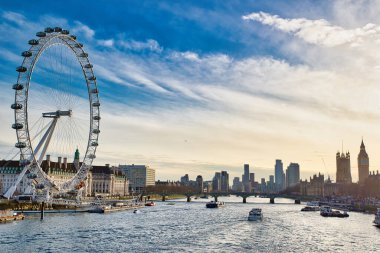 London Eye, Thames nehri, birkaç tekne ve parçalı bulutlu gökyüzünün altındaki parlamento binalarının yer aldığı gökyüzü..