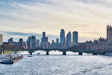 Westminster Köprüsü, yüksek binalar, Parlamento Binaları ve birkaç tekne de dahil olmak üzere çok sayıda binanın yer aldığı Thames Nehri 'nin karşısındaki Londra şehir manzarası. Gökyüzü çoğunlukla bulutlu ve solgun bir gün batımı var..
