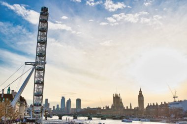 London Eye, Houses of Parliament, Big Ben ve parçalı bulutlu gökyüzünün altındaki Thames Nehri 'nin bir kısmının yer aldığı Londra silueti. Binalar, tekneler ve bir vinç de görülebilir durumda..