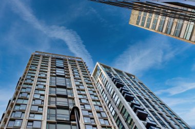 İnce bulutlu, parlak mavi gökyüzüne karşı üç modern gökdelenin alçak açılı görüntüsü. Bu binalar öncelikle Londra, İngiltere 'de koyu renk pencere çerçeveli açık bej ve beyazdır..