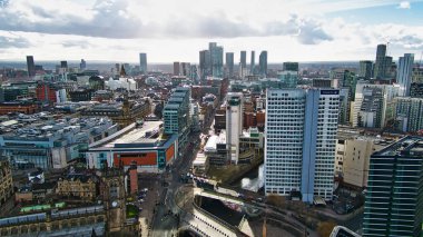 Manchester şehir merkezinin yüksek açılı görüntüsü modern ve eski binaların, gökdelenlerin ve kanalların yoğun bir karışımını sergiliyor. Görüntü, Manchester, İngiltere 'de parçalı bulutlu bir gökyüzü altında mimari tarzların ve çeşitli yapı yüksekliklerinin bir karışımını içerir..