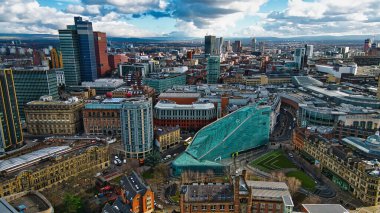 Manchester şehir merkezinin hava görüntüsü, gökdelenler ve belirgin yeşil-mavi kaplama bina da dahil olmak üzere modern ve tarihi mimarinin bir karışımını sergiliyor. Parçalı bulutlu bir gökyüzünün altında genişleyen şehir manzarası.
