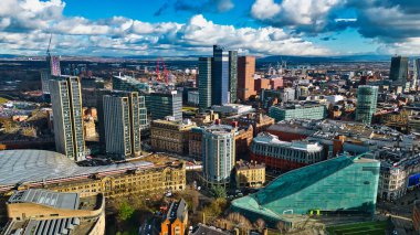 Manchester şehir merkezinin hava görüntüsü, çok sayıda gökdelen, modern ve tarihi binalar ve parçalı bulutlu bir gökyüzü altında bir tren istasyonu sergileniyor..