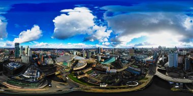 Manchester, İngiltere 'nin panoramik hava görüntüsü, parçalı bulutlu bir gökyüzü altında şehir manzarasını gösteriyor. Önde gelen stadyum ve modern yüksek binalarla sayısız bina, yol ve altyapı görülebilir durumda..