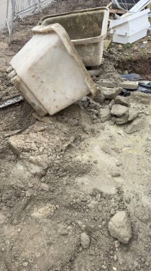 Kazılmış toprak ve molozların arasında iki adet beyaz plastik yapı küveti bulunuyor. Bir küvet ters dönmüş ve içindekiler dökülmüş. Zemin gevşek toprak, taşlar ve çamurdan oluşuyor..