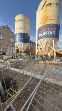 Two large Cemex mortar silos stand on a construction site.  A damaged metal barrier and an excavated trench with yellow pipe are in the foreground.  A partially-visible house is in the background. clipart