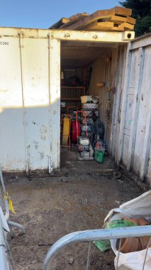 Kısmen açık olan beyaz bir nakliye konteynırı çeşitli inşaat ekipmanı ve malzemelerini ortaya çıkarıyor. Konteynırı çamurlu zemin kaplar; kil borular ve bir uyarı bandı ön planda görünür. Tahta kalaslar konteynırın üzerinde duruyor..