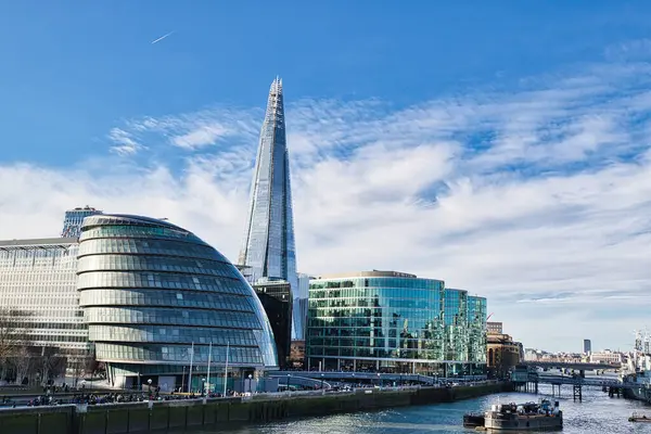 Nehir boyunca Çömlek, Belediye Binası ve modern cam binaların yer aldığı Londra silueti. Parçalı bulutlu mavi bir gökyüzü parlak bir zemin sağlar. Nehir kıyısında insanlar görülebilir..