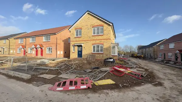 Yeni inşa edilmiş tuğladan evler geliştirme aşamasında. Peyzaj düzenleme tamamlanmamış, inşaat malzemeleri ve enkaz parçaları görülebiliyor. Ana odak noktası iki katlı sarı tuğlalı ev..