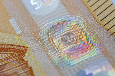 Banknotun güvenlik özelliğinin yakın plan görüntüsü; notun tasarımının bir kısmına geometrik desenli yanardöner folyo. Arkaplanda kahverengi ve bej renkler hakim.