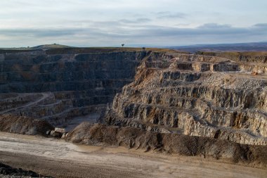 Açıkta kaya tabakası olan büyük, teraslı bir taş ocağının havadan görünüşü. Çamurlu yollar ve toprak yığınları görülebilir. Ağır makineler mevcut.
