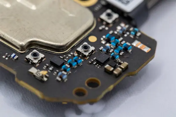 Bir devre kartı parçasının yakın plan görüntüsü, metalik elementler, yüzey dayanıklıları ve küçük düğmeler de dahil olmak üzere çeşitli bileşenleri gösteriyor. Ağırlıklı olarak koyu tonlarda metalik gri ve mavi aksanlı..