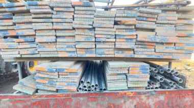 Paslı kırmızı bir platforma yığılmış iskeleler ve metal borular. Kalaslar aşınma ve çeşitli boya izleri gösteriyor. Borular paketlenmiş, uçları görünür..