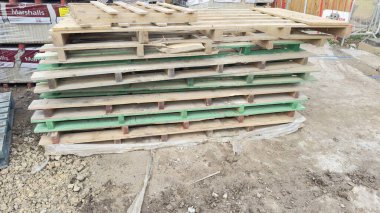 Bir yığın aşınmış ahşap paletler, bazıları yeşil boyalı bölümler, çıplak zemin üzerinde yatıyor. Birkaç palet hasar görmüş. Arkaplanda inşaat malzemeleri yer alıyor.