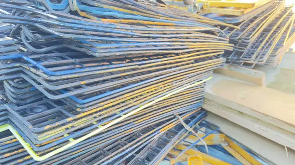 Büyük bir yığın yıpranmış, mavi ve sarı metal ızgaralar, muhtemelen inşaat şebekesi, dışarıda ahşap kalasların yanında duruyor. Şebeke aşınma ve pas belirtileri gösteriyor..