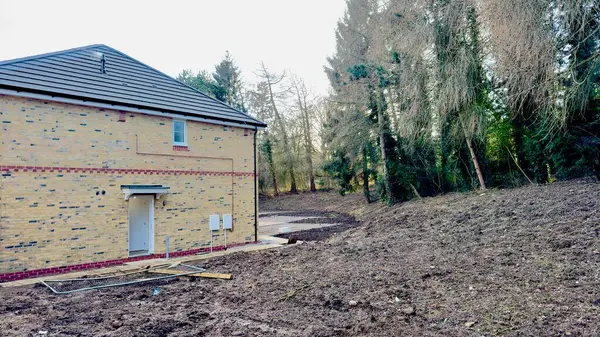 Yeni yapılmış, koyu gri çatılı açık sarı tuğlalı bir evin arka manzarası. Ev, temiz arazi ve yaprak döken ağaçların yanında yer alıyor. Toprak çıplak ve bazı kalıntılar görülebilir..