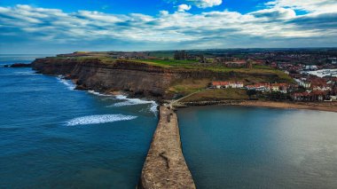 Kayalıkların, binaların ve Whitby, İngiltere 'de dağınık bulutların olduğu mavi gökyüzünün bulunduğu bir kıyı şeridine uzanan yüksek açılı bir iskele..