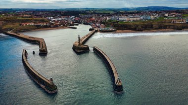 Kasabayı çevreleyen üç dalgalı limanın havadan görüntüsü. Bir deniz feneri bir dalgakıranın üzerinde duruyor. İngiltere, Whitby 'de gri gökyüzü ve okyanus.