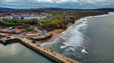 Bir rıhtım, sahil, uçurum ve İngiltere 'nin Whitby şehrinde bulutlu bir gökyüzü altında bir dizi binanın yer aldığı bir kıyı kasabasının havadan görünüşü..