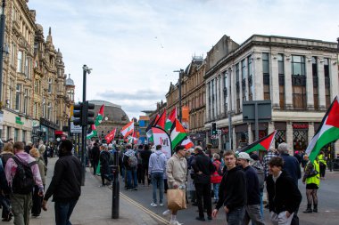Bir şehir caddesinde bayraklarla yürüyen kalabalık, yan taraflarda binalar var. Bulutlu gökyüzü. Ağırlıklı olarak sessiz renkler.