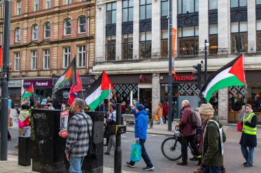 Sokak düzeyinde bir gösteri, arka planda binalar ve dükkanlar bulunan bir şehir caddesinde Filistin bayrakları tutan insanların yer aldığı.