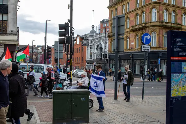 Filistin ve İsrail bayrakları sergileyen insanların yürüdüğü bir sokak sahnesi. Binalar ve trafik ışıkları görülebilir.