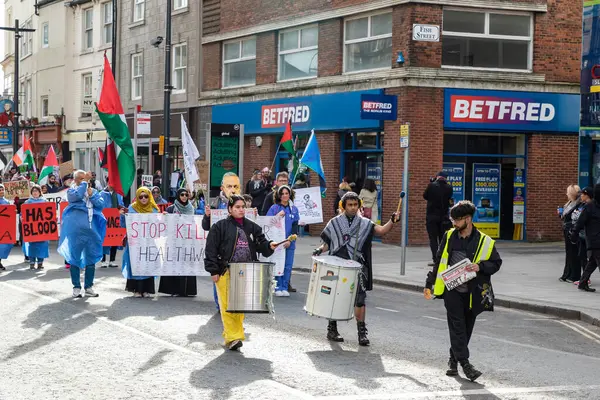 Şehir caddesinde binalar ve bir Betfred spor kitabı ile hareket eden tabelalar, bayraklar ve davullarla gündüz protesto yürüyüşünün sokak seviyesinde görüntüsü.