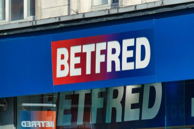 Görüntü, Manchester, İngiltere 'deki mağaza vitrinine yansıyan kırmızı dikdörtgen ve mavi arkaplan üzerinde koyu beyaz harflerle Betfred şirketinin logosunu gösteriyor..