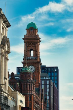 Yeşil kubbeli, kısmen diğer binalar tarafından gizlenmiş, Manchester, İngiltere 'de bulutlu mavi gökyüzüne bakan tuğladan bir saat kulesi görülüyor..